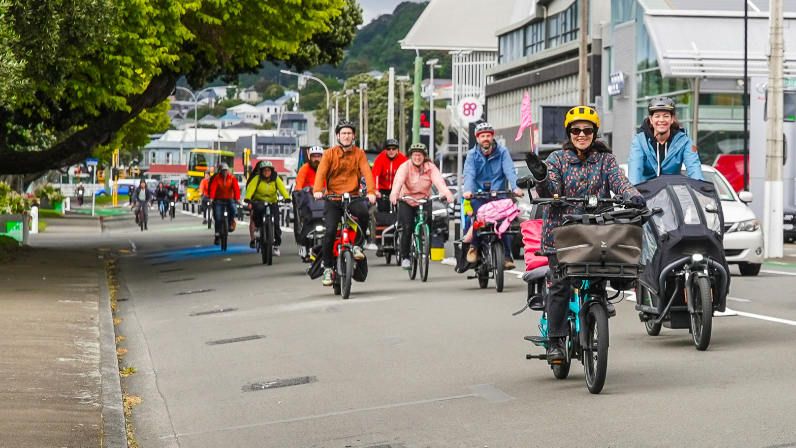 Group of people riding bicycles and electric bikes on a city street with trees and buildings in the background.